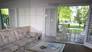 A living room with a sofa, a coffee table with remote controls and magazines, and a sliding glass door leading to an outdoor patio with chairs and a table.