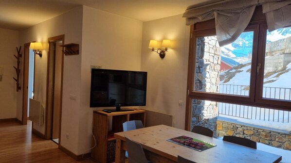 A room with a wooden table, a flat-screen TV, two wall-mounted lamps, and a window with a view of a stone building and mountains.