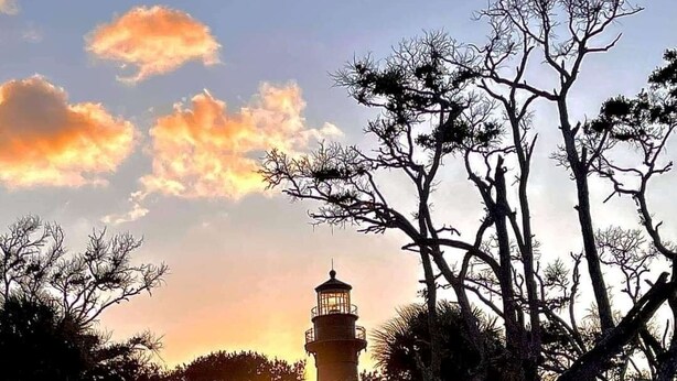 Ein Leuchtturm mit freiem Blick auf den Sonnenuntergang, umgeben von Bäumen und einer Sandfläche.