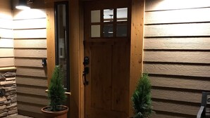 A wooden door with a glass panel, flanked by two potted plants, leading to a brick-paved entrance.