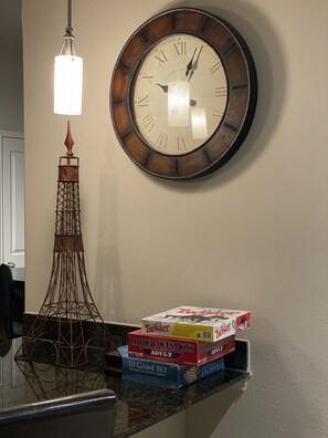 A clock with Roman numerals and a pendulum hangs above the kitchen counter, accompanied by a decorative metal tower and a selection of board games.