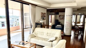 A spacious living room with a large sliding glass door leading to a balcony, a white sofa, a glass coffee table, and a wooden floor. - Minpaku Arin Kurin (Haebaru)