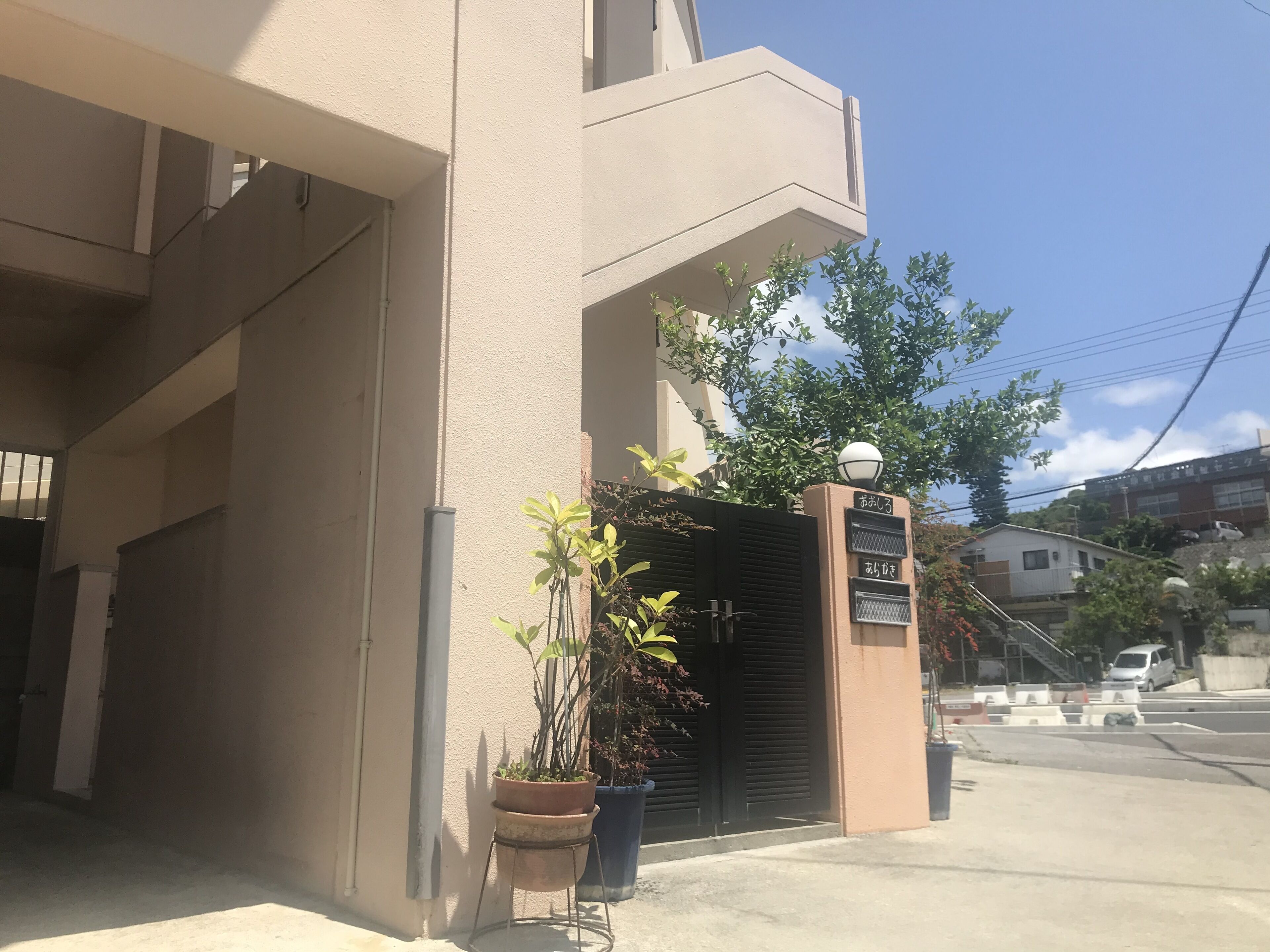 Un edificio con facciata color beige, un cancello nero e un cartello con caratteri giapponesi. Davanti al cancello ci sono piante in vaso.