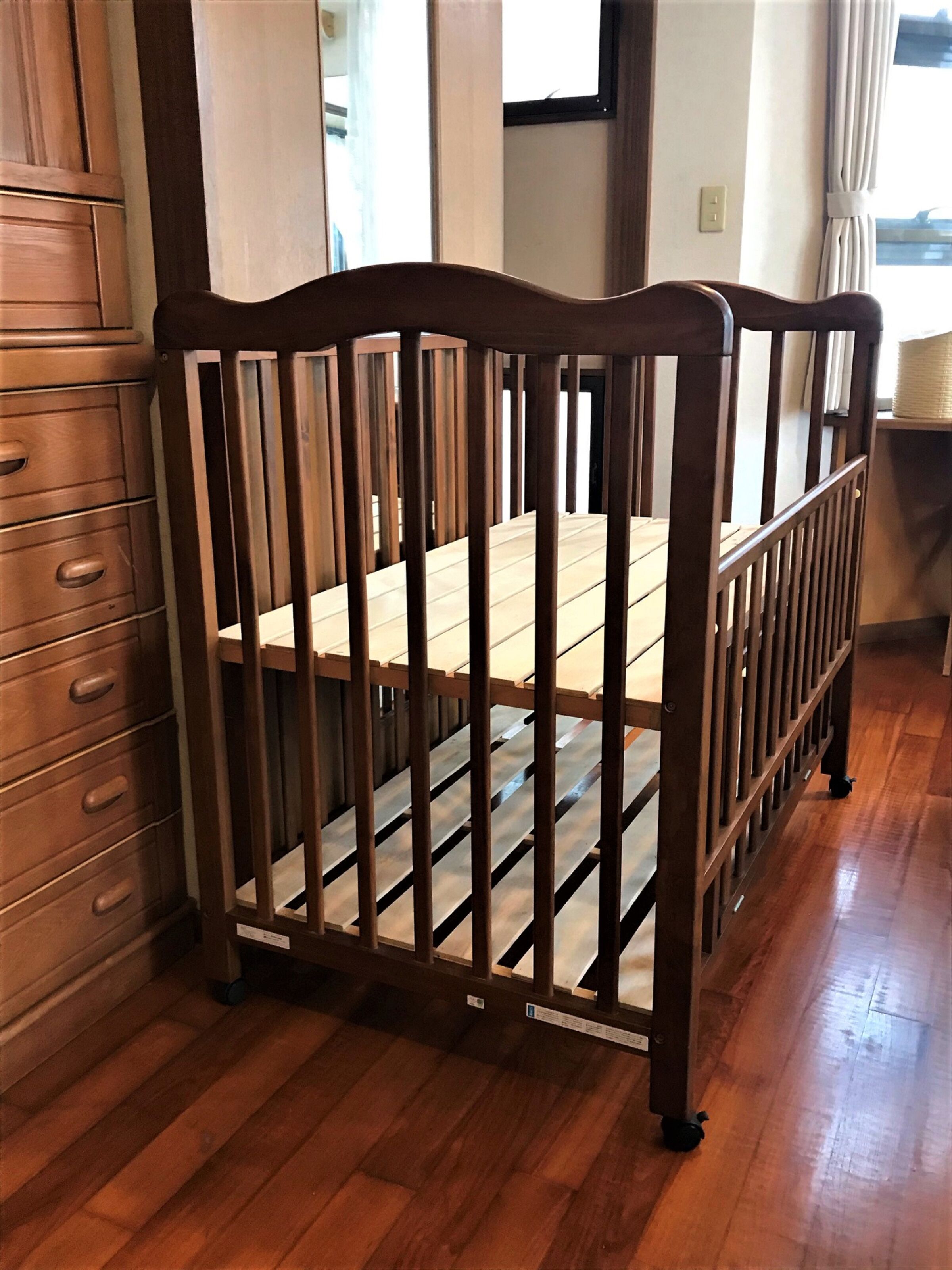 A wooden crib with a slatted design, positioned in a room with wooden flooring and a dresser to the left.