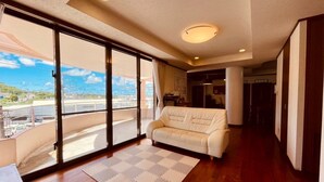 A spacious living room with a large sliding glass door leading to a balcony, a white sofa, and a wooden floor. - Minpaku Arin Kurin (Haebaru)