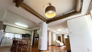 A spacious living area with a wooden dining table and chairs, a large light fixture, and a kitchen in the background. - Minpaku Arin Kurin (Haebaru)