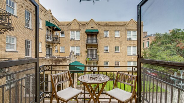 Un balcón con dos sillas de madera y una mesa redonda pequeña, con vista a un edificio de ladrillo con múltiples ventanas y balcones.
