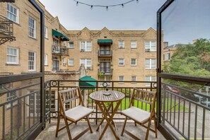 Un balcon avec deux chaises en bois et une petite table ronde, donnant sur un immeuble en briques doté de nombreuses fenêtres et de balcons.