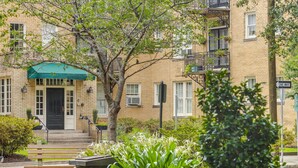 Un edificio de varios pisos con un toldo verde sobre la entrada, un árbol en la parte delantera y una señal de tráfico que indica 'ONE WAY'.
