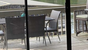 Une terrasse dotée de chaises et d’une table en osier, offrant une vue sur un lac, avec une zone herbeuse et des arbres en arrière-plan.