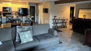Un salon moderne avec un canapé gris, un ventilateur de plafond et un coin cuisine à l’arrière-plan.