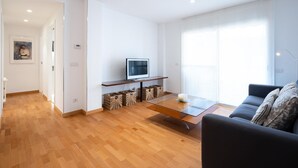 A modern living room with a wooden floor, a glass coffee table, a black sofa, a TV on a wooden stand, and a large window. - Spacious apartment with A/C and heating (L'Ametlla de Mar)