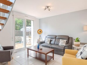 A modern living room with a grey sofa, a yellow armchair, and a wooden coffee table. There is a large window with a view of the outside, and a ceiling light fixture above the sofa.