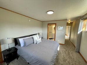 Une chambre avec un grand lit, une table de chevet, une lampe, un luminaire au plafond, une porte donnant sur une salle de bain et un tableau accroché au mur.