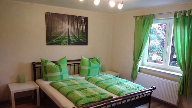 A bedroom with a bed, bedside tables, a window with curtains, a radiator, and a forest-themed wall hanging. - Lower vacation apartment between Tauber Valley, Odenwald and Bauland (Ahorn)
