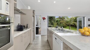 A modern kitchen with a central island, built-in appliances, and a view of the garden. - Pohutukawa Peace - Mangawhai Holiday Home (Mangawhai Village)