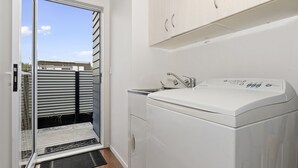 A compact laundry room with a washing machine, a sink, and a sliding glass door leading to a balcony. - Pohutukawa Peace - Mangawhai Holiday Home (Mangawhai Village)
