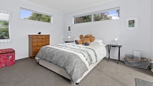 A bedroom with a large bed, a wooden dresser, a desk with a lamp, and a red suitcase. - Pohutukawa Peace - Mangawhai Holiday Home (Mangawhai Village)