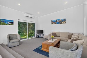 A modern living room with a sofa, armchair, and a coffee table. There is a television and a bookshelf with books and DVDs.