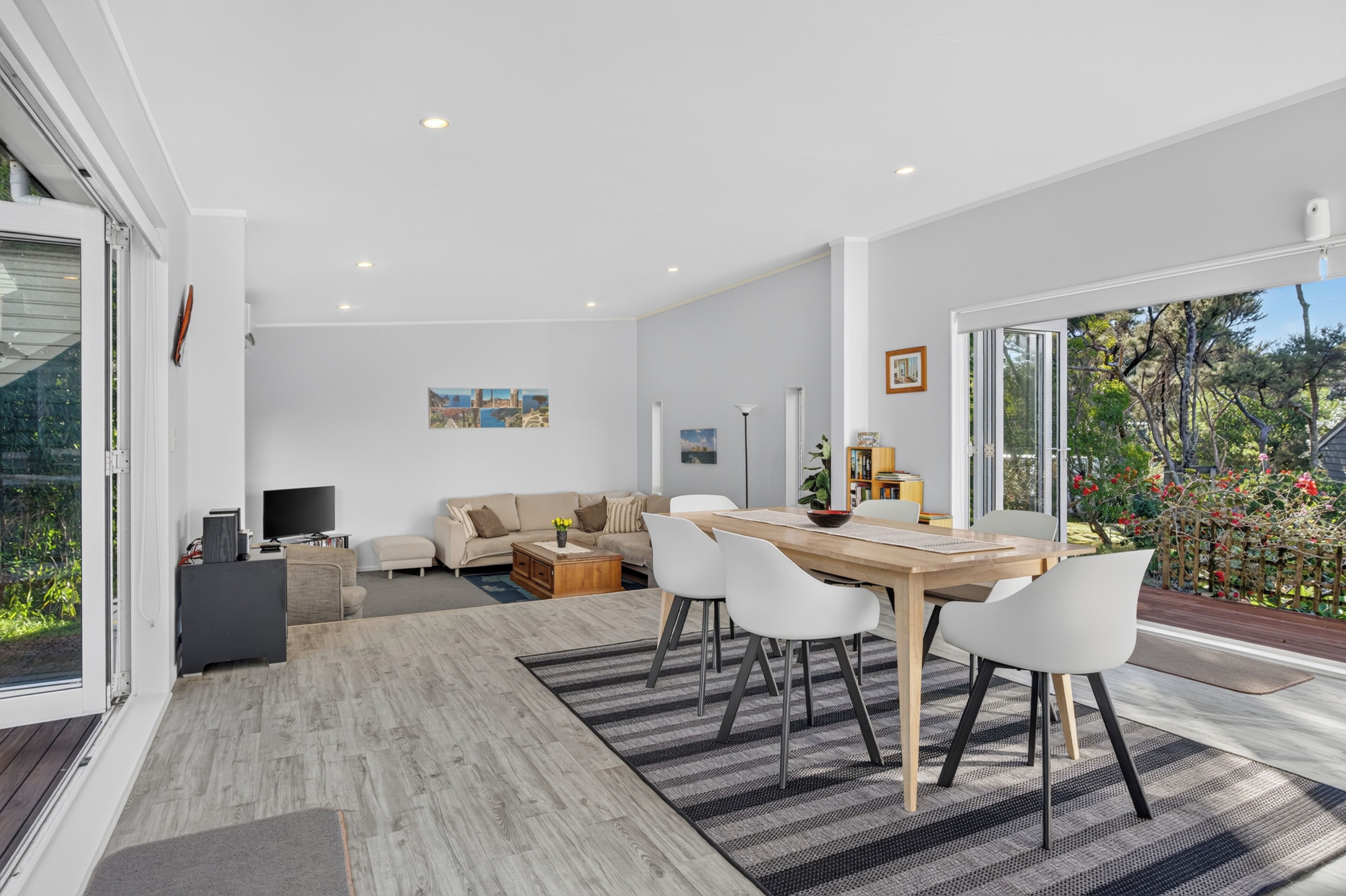 Un salon moderne avec un canapé, une table basse et un téléviseur. Il y a également un coin repas avec une table et des chaises. La pièce est dotée de grandes fenêtres et offre une vue sur la verdure environnante.
