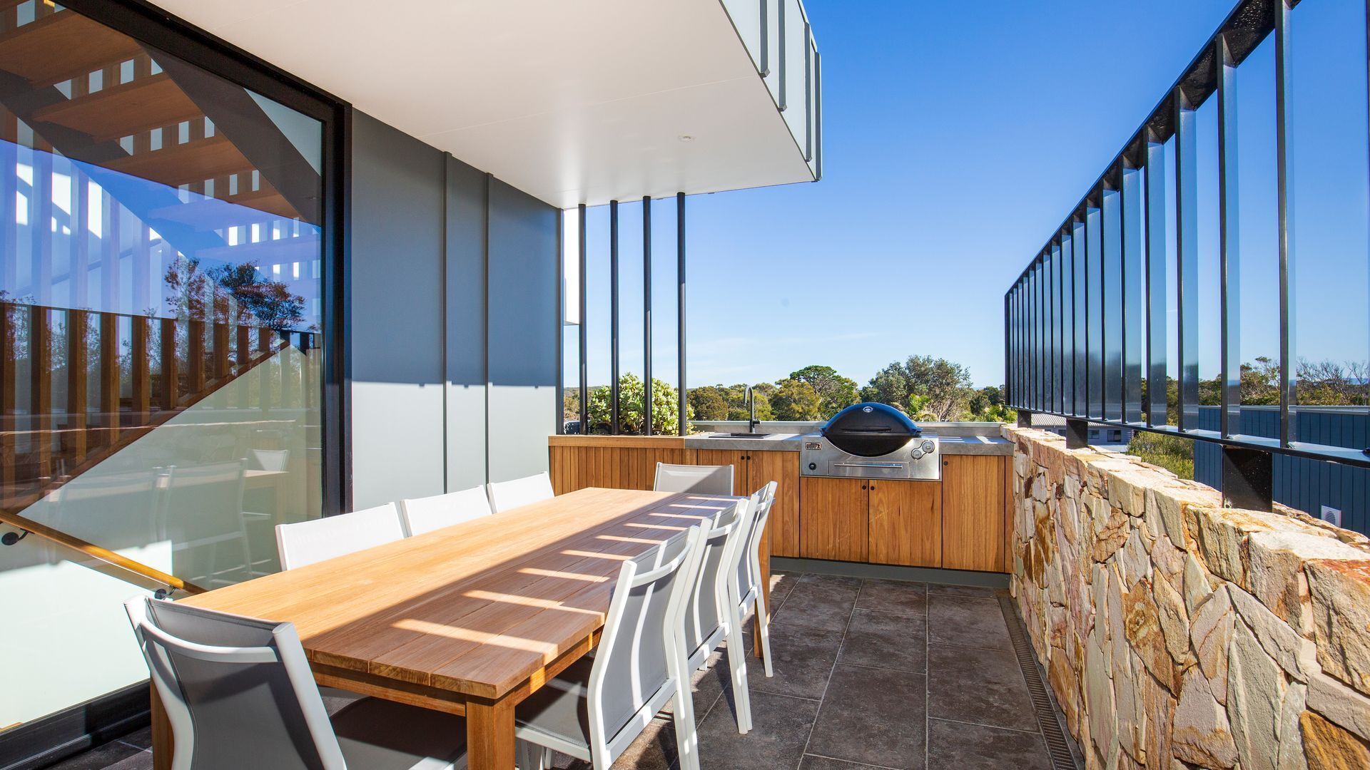 A modern outdoor dining area with a wooden table and chairs, a barbecue grill, and a stone wall.