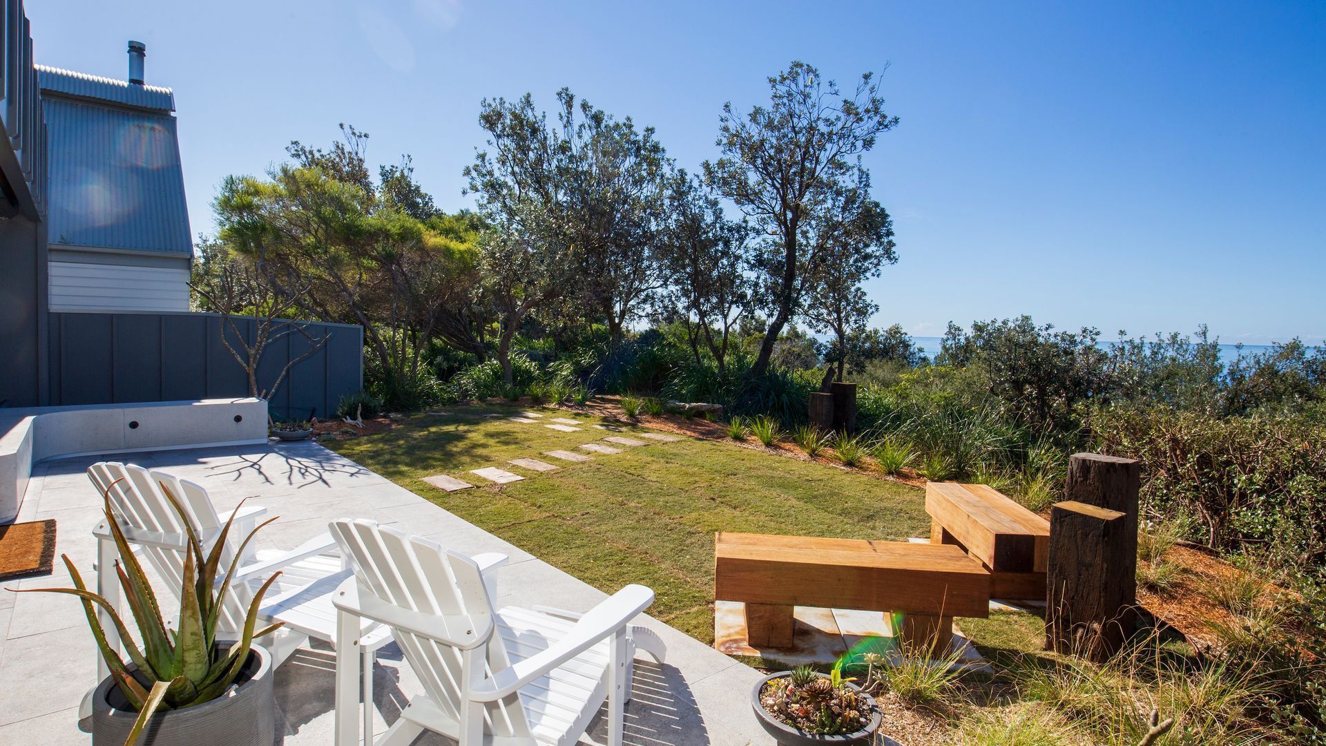A patio with white chairs and a wooden bench, surrounded by a garden with various plants and trees, leading to a view of the sea.