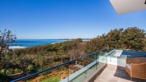 Ein Balkon mit Glastrennwand, der auf eine Küstenlandschaft mit Bäumen und dem Meer blickt.
