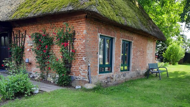Une maison de campagne en briques, avec un toit de chaume, un petit jardin parsemé de fleurs rouges et un banc dans la cour.