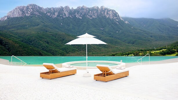 Ein Poolbereich mit Liegestühlen und einem Sonnenschirm, mit Blick auf ein türkisfarbenes Schwimmbecken und eine gebirgige Landschaft.