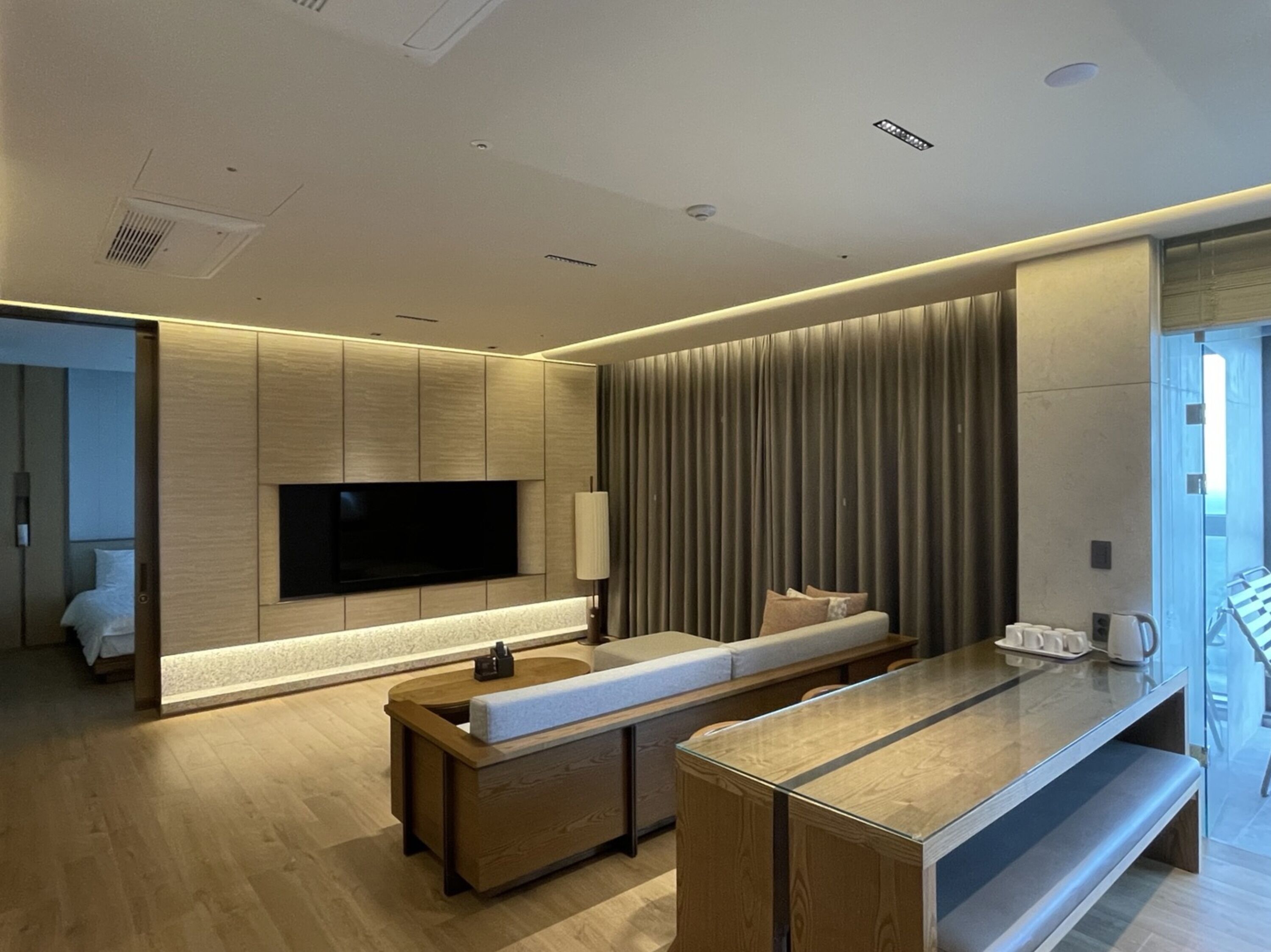 A modern hotel room with a large flat-screen TV mounted on a wooden panel wall, a seating area with a bench and a coffee table, and a large window with curtains.