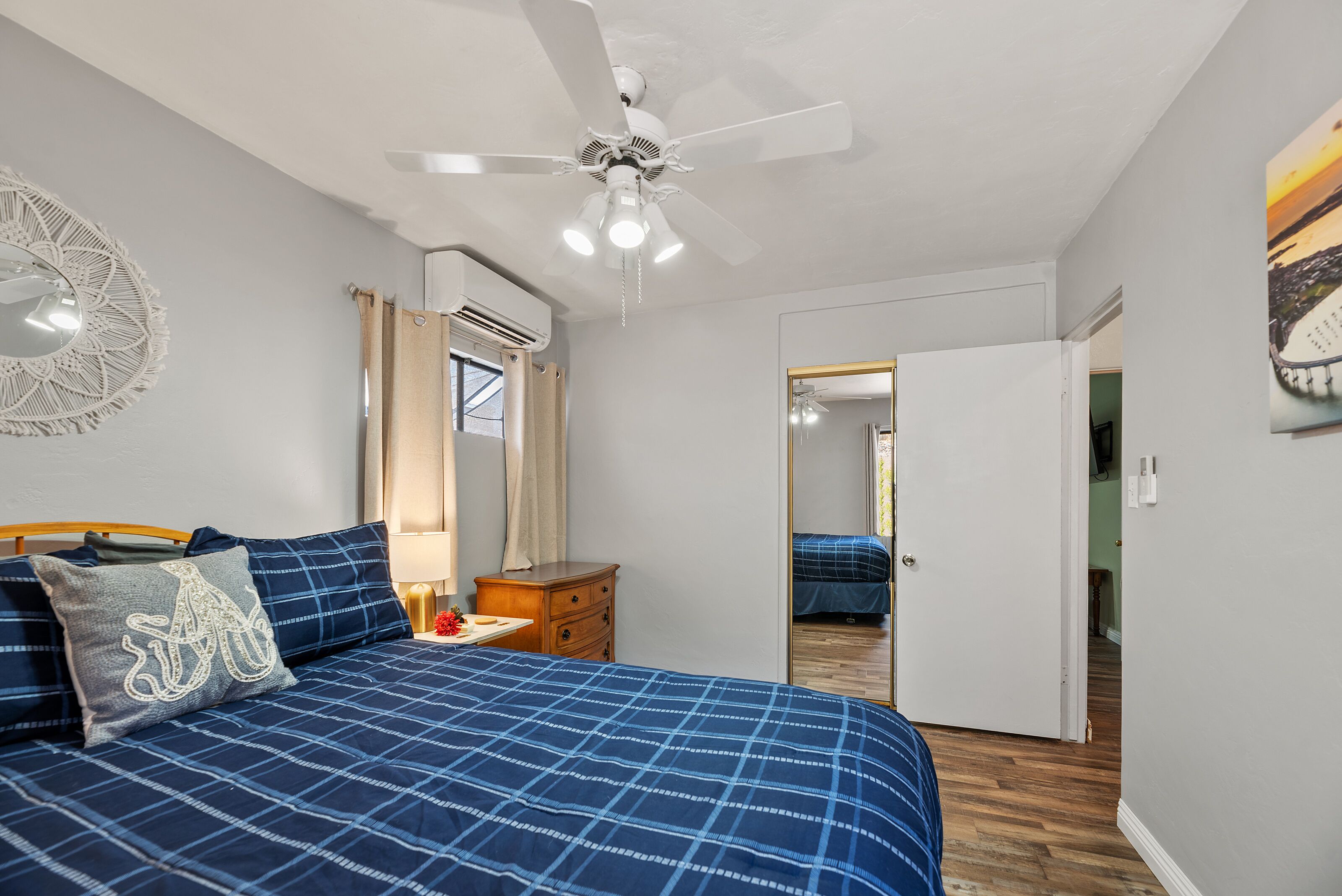 A bedroom with a bed, a ceiling fan, an air conditioning unit, a mirror, and a door leading to another room.