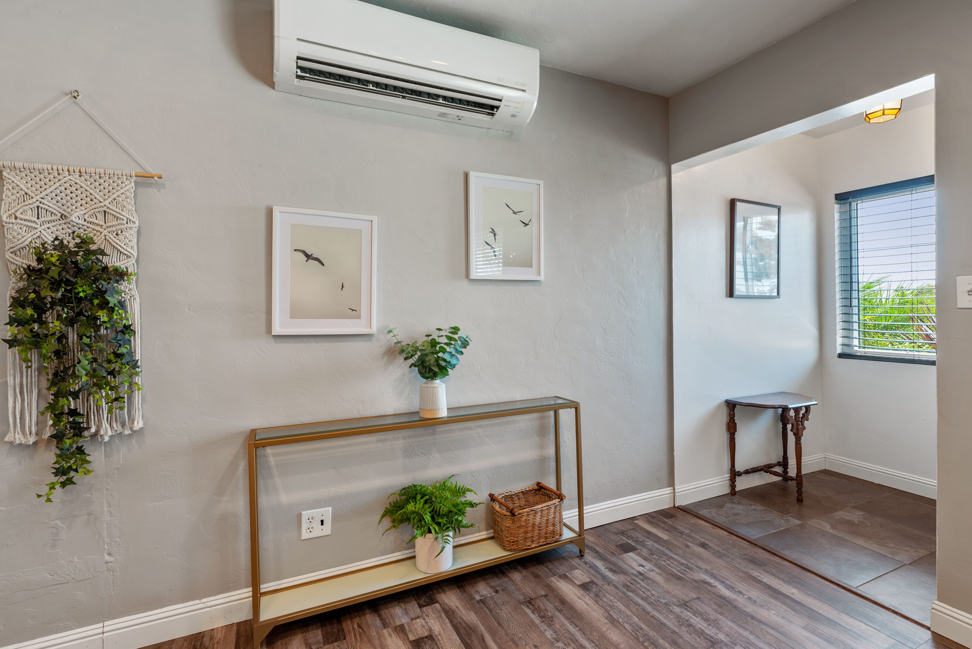 Un couloir doté d’un plancher en bois, d’un climatiseur fixé au mur, d’une décoration murale en macramé, d’œuvres d’art encadrées, d’une console sur laquelle sont disposées des plantes et un panier, et d’une fenêtre avec des stores.