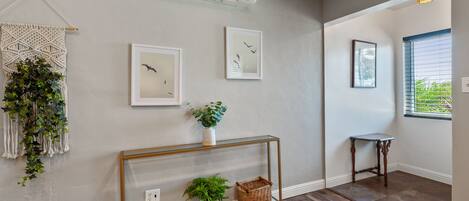Un couloir doté d’un plancher en bois, d’un climatiseur fixé au mur, d’une décoration murale en macramé, d’œuvres d’art encadrées, d’une console sur laquelle sont disposées des plantes et un panier, et d’une fenêtre avec des stores.
