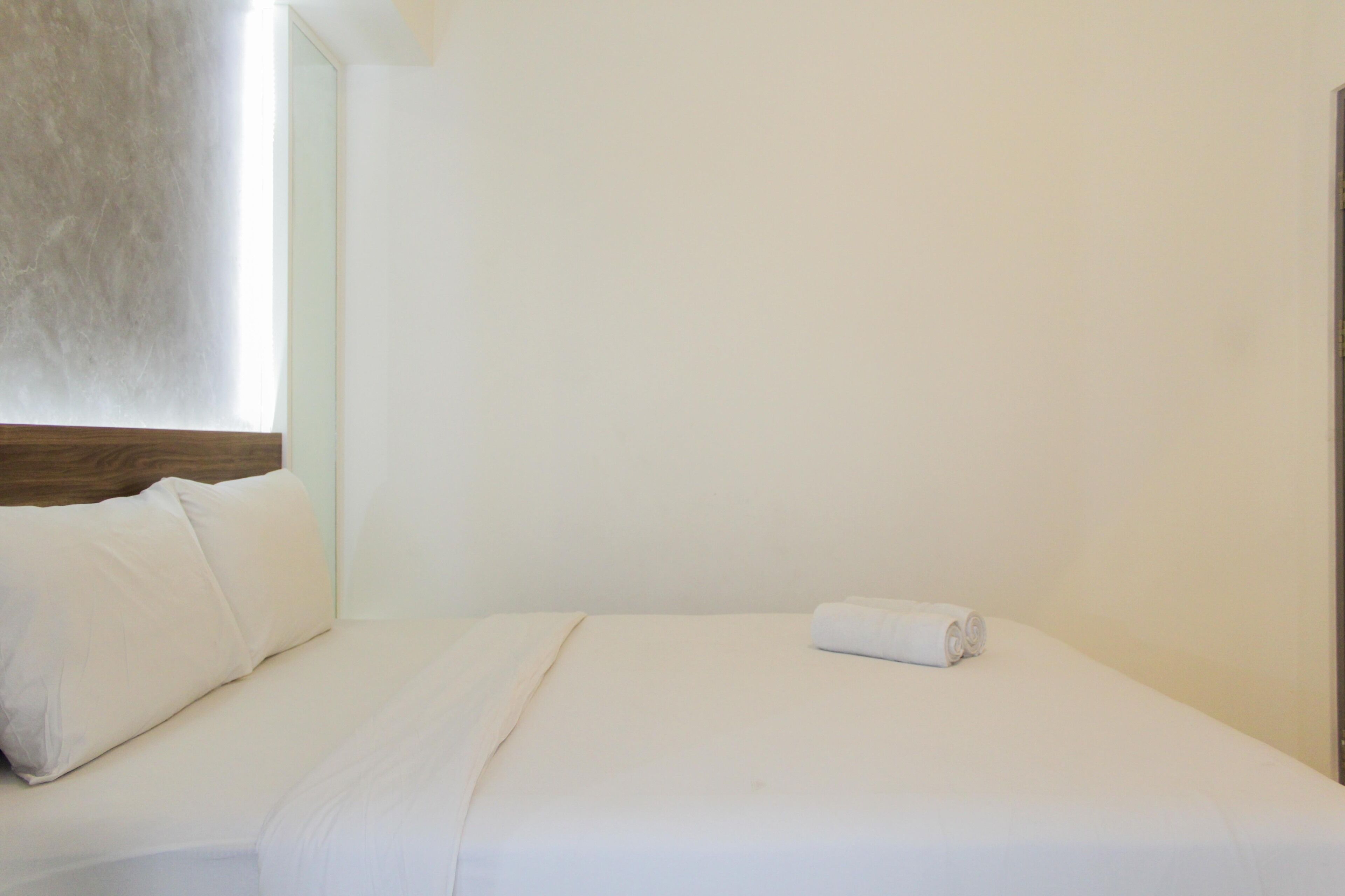 A neatly made bed with white linens and a rolled towel, a wooden headboard, and a plain white wall.