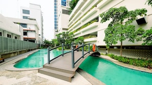 Ein moderner, überdachter Pool mit Holzdeck und Metallgeländern, umgeben von viel Grün und angrenzend an ein Mehrstöckiges Gebäude mit Balkonen.