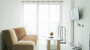 Ein modernes Wohnzimmer mit einem beigen Sofa, einem kleinen Tisch mit Blumenvase, einem an der Wand montierten Fernseher und einem Balkon mit weißen Vorhängen.
