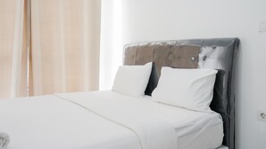 Ein ordentlich bezogenes Bett mit weißen Leinen und einem grauen Kopfteil in einem Zimmer mit schlichter weißer Wand und einem Fenster mit beigen Vorhängen.