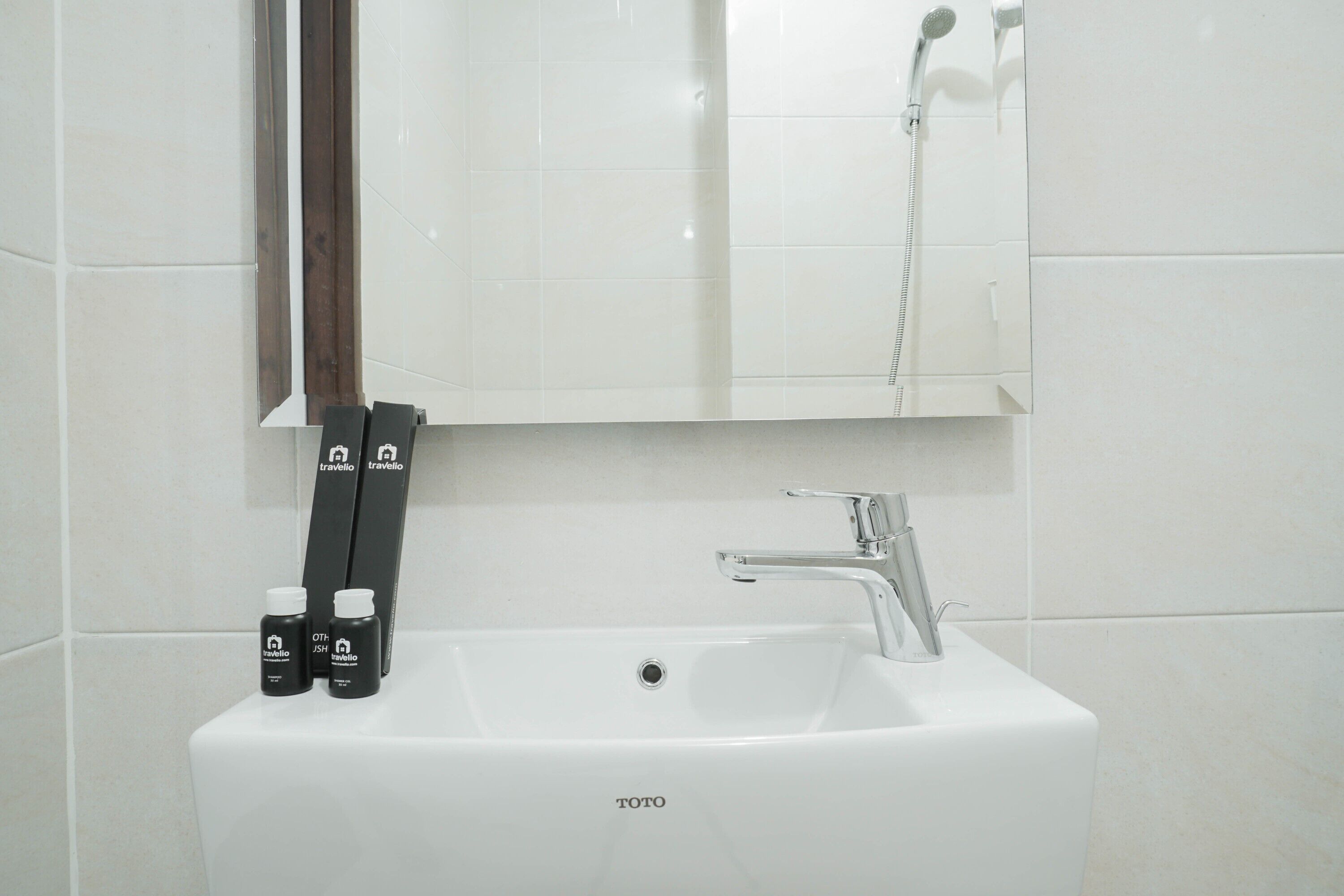Un lavabo blanc dans une salle de bain, avec un miroir au-dessus, une pomme de douche et deux flacons de produits de toilette.