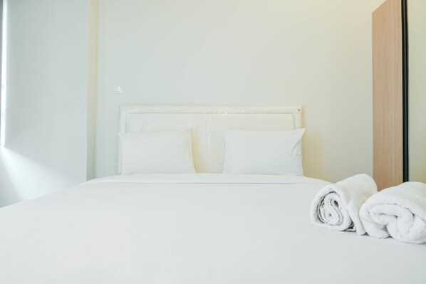 A neatly made bed with white linens and pillows, a wooden headboard, and two rolled towels on the side.