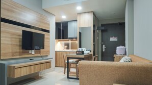 A modern living room with a flat-screen TV, wooden furniture, a dining area, and a kitchen in the background. - Elegant 1BR Apartment with Working Space Mustika Golf Residence (Cikarang)