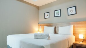 A neatly made bed with white linens and a folded towel, a wooden headboard, and a nightstand with a lamp. - Elegant 1BR Apartment with Working Space Mustika Golf Residence (Cikarang)
