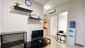 Una sala de estar moderna con un televisor de pantalla plana, una mesa de centro, un mueble negro para la televisión, un dispensador de agua y una pequeña cocina con estufa y armarios.