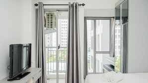 Hotellihuone, jossa on sänky, jalustalla oleva televisio, parveke, jossa on verhot, ja ikkuna, josta on näkymä rakennuksiin.