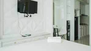 A modern hotel room with a flat-screen TV mounted on the wall, a white vanity with drawers, a small table with a chair, and a kitchen area with cabinets and a refrigerator.