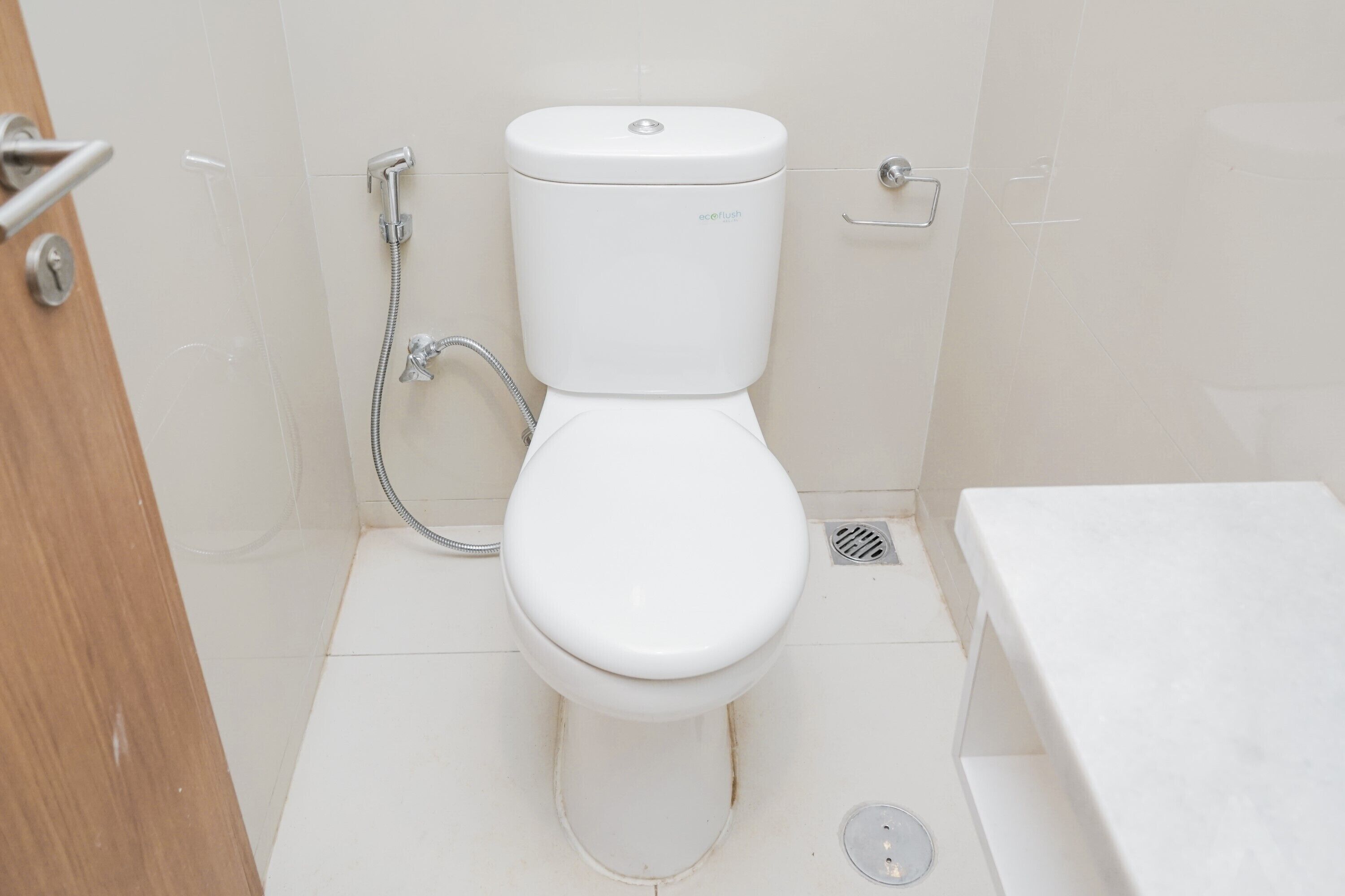 Des toilettes blanches, équipées d’un pommeau de douche, dans une salle de bain aux carreaux blancs.