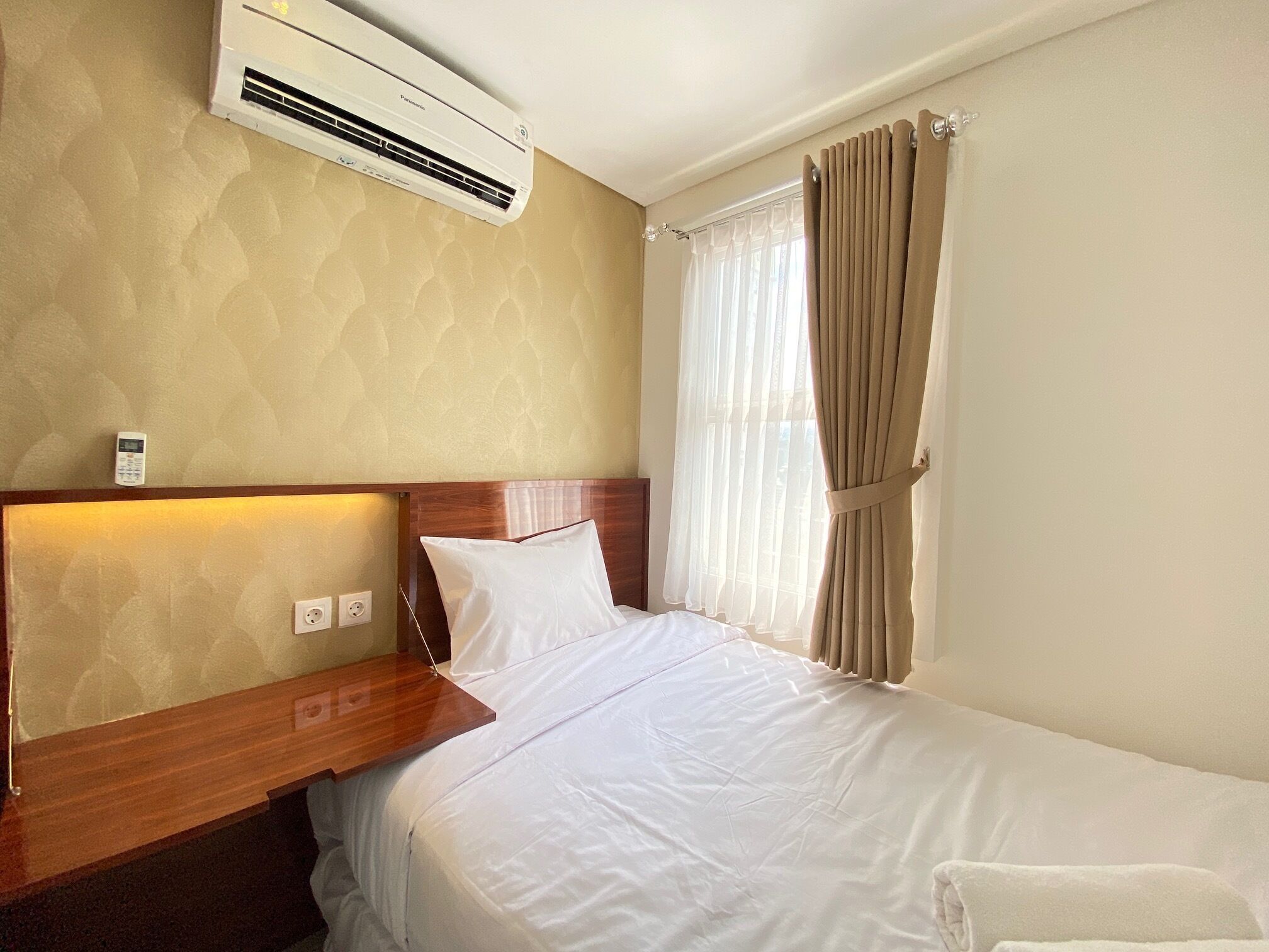 Ein Hotelzimmer mit Bett, Nachttisch, Klimaanlage, einem Fenster mit Vorhängen und einem wandmontierten Thermostat.
