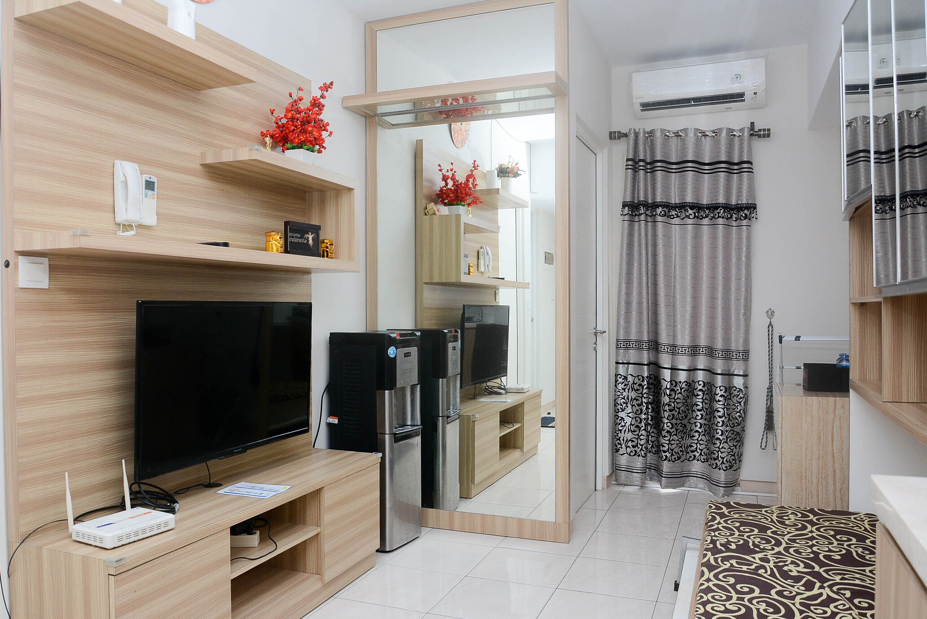 A modern living room with a flat-screen TV, a refrigerator, and a coffee machine. There is a wooden shelf with decorative items and a mirror reflecting the room. A patterned rug is on the floor, and a curtain is visible in the background.