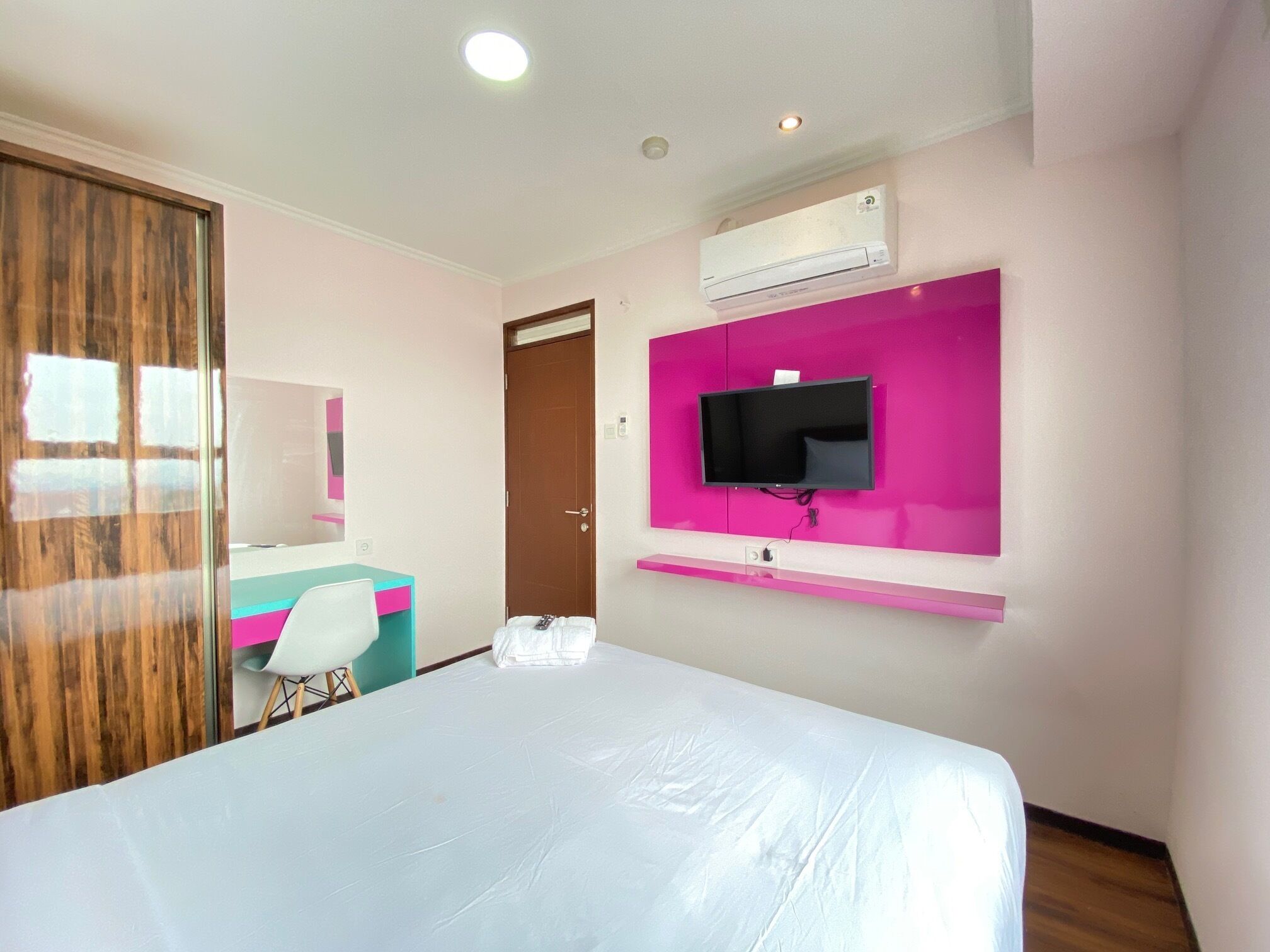 Habitació d'hotel amb un llit, un televisor fixat a una paret rosa, un aparell d'aire condicionat i una porta de fusta.