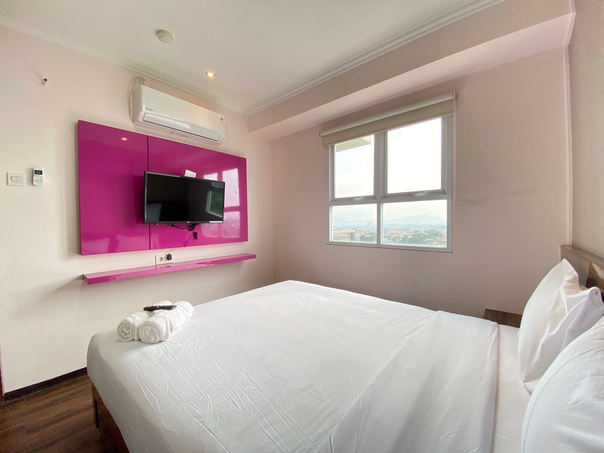 Quarto de hotel com uma cama, uma TV fixada em uma parede rosa, uma janela com vista para a cidade e um aparelho de ar condicionado.