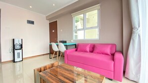 A modern living room with a wooden coffee table, a pink sofa, a dining area with a table and chairs, and a refrigerator. - Modern & Cozy 2BR at Gateway Pasteur Apartment (Cimahi)